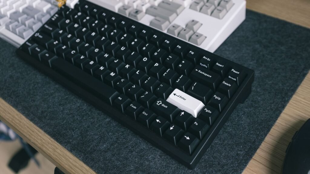 Black compact mechanical keyboard with a white Enter key on a gray felt desk mat, with a white keyboard behind it