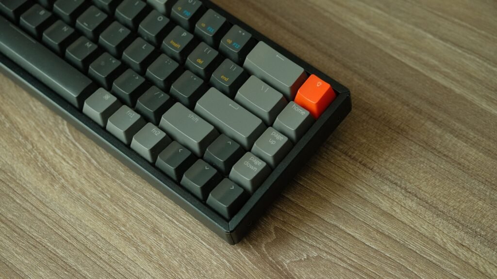 Close-up of the right side of a compact dark gray mechanical keyboard with an orange accent key in the top corner, on a wooden table