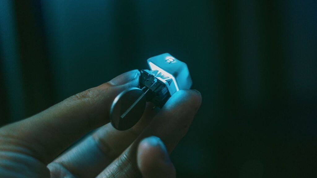 A hand holding a single backlit keycap marked R, still attached to its mechanical switch with the metal pins visible underneath