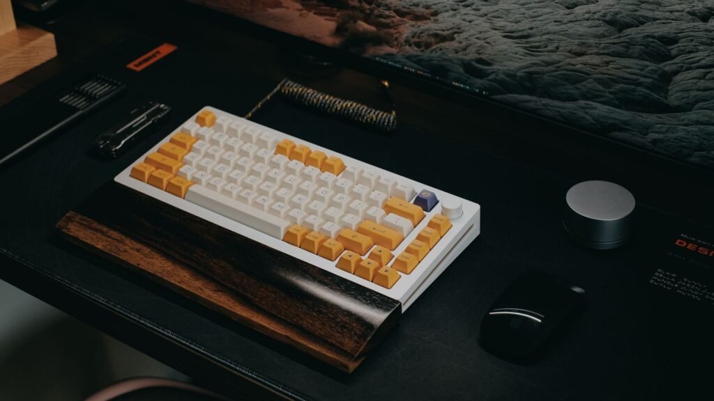 White 75 percent mechanical keyboard with yellow accent keycaps and a volume knob, resting against a dark wooden wrist rest on a black desk mat