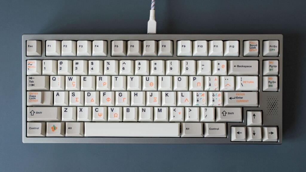 Overhead view of a gray compact mechanical keyboard with cream keycaps, a function row, arrow keys and a white USB-C cable on a blue surface