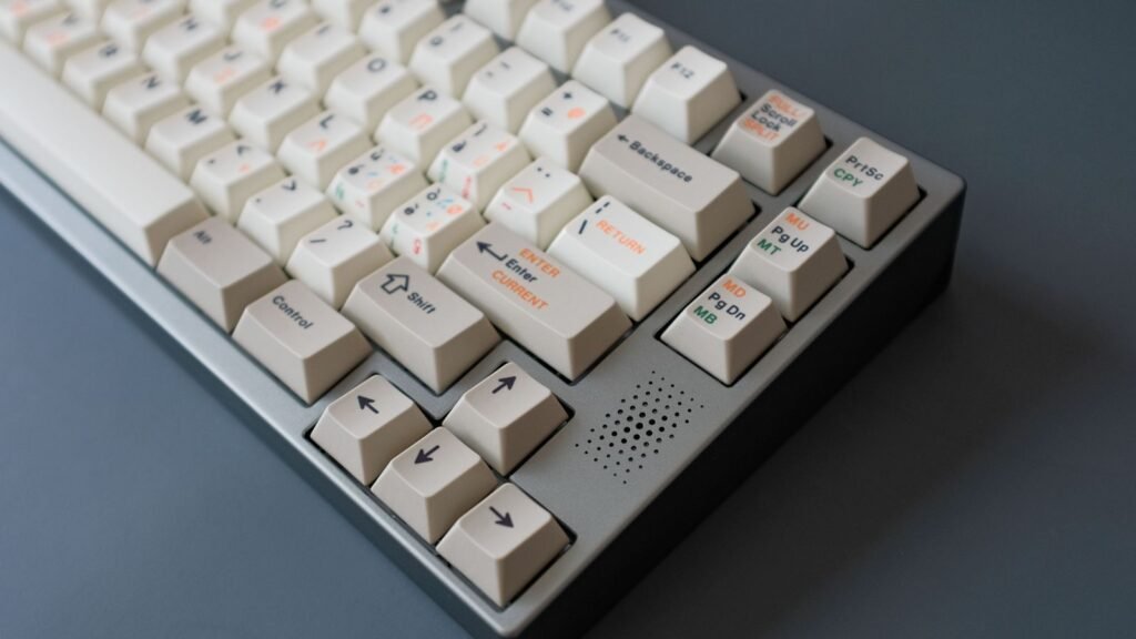 Close-up of a mechanical keyboard with cream keycaps printed in orange, gray and green legends, sitting in a gray metal case with a speaker-style grille next to the arrow keys