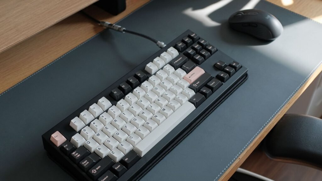 Black tenkeyless mechanical keyboard with white and dark keycaps and a pink Escape key, connected by a coiled cable, on a gray desk mat next to a black mouse