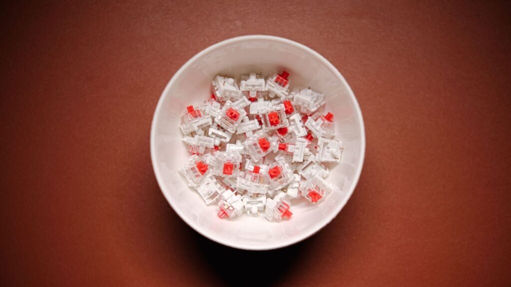 Top view of a white bowl filled with loose mechanical keyboard switches with clear housings and red stems, on a brown surface