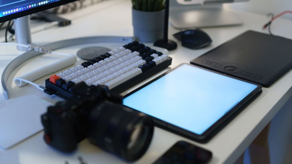 A black and white tenkeyless mechanical keyboard with a coiled white cable on a white desk next to a tablet, mouse and camera