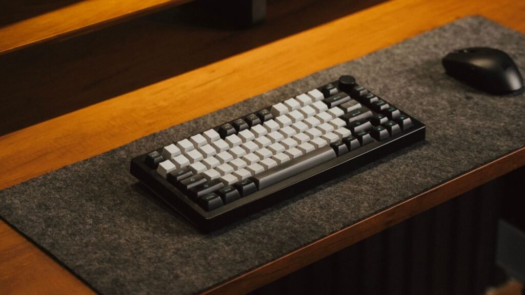 A black 75% mechanical keyboard with white and dark gray keycaps and a rotary knob on a gray felt desk mat beside a black mouse