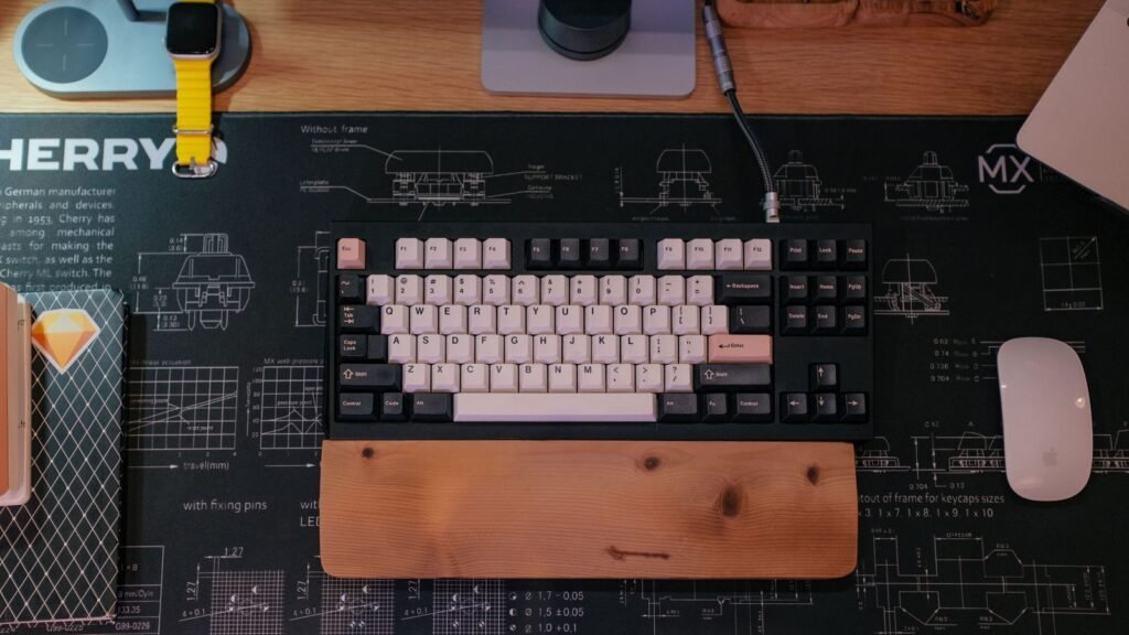 Overhead view of a tenkeyless mechanical keyboard with cream and dark gray keycaps, a wooden wrist rest and a white mouse on a desk mat printed with switch diagrams
