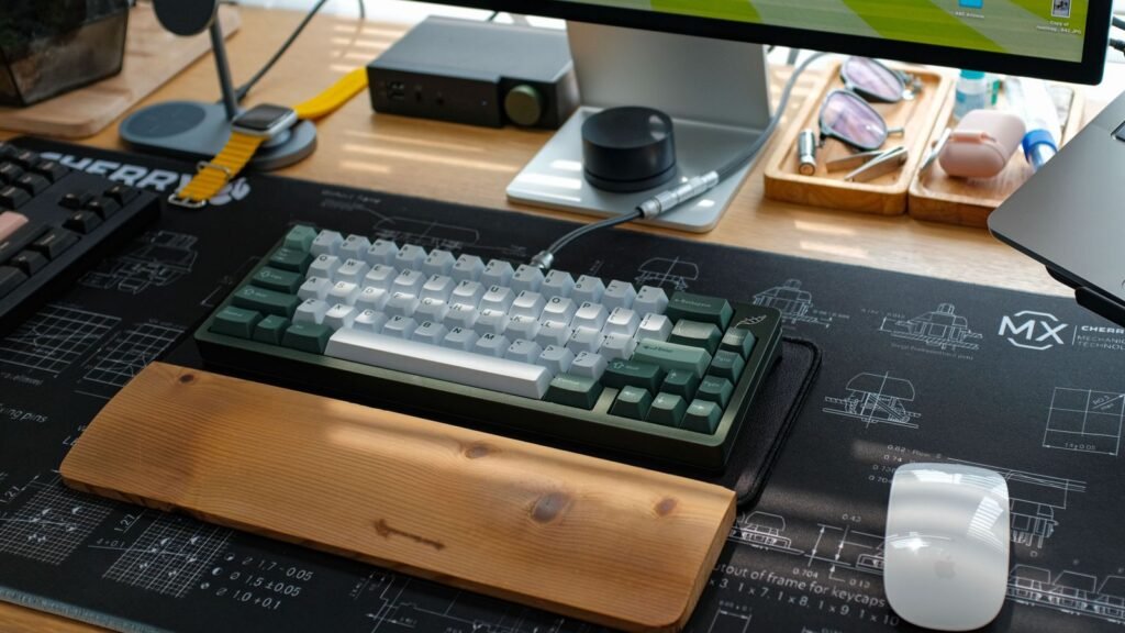 A green and white compact mechanical keyboard with a wooden wrist rest on a large desk mat, next to a white Apple Magic Mouse, a monitor and the edge of a laptop