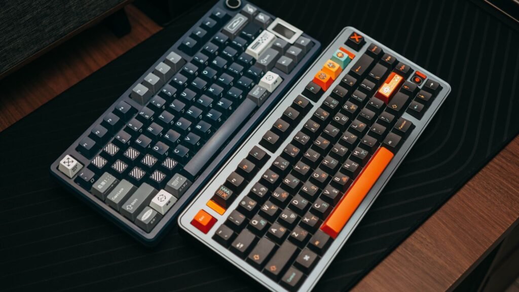 Two custom mechanical keyboards side by side on a black desk mat, one navy with a rotary knob and one silver with dark and orange keycaps