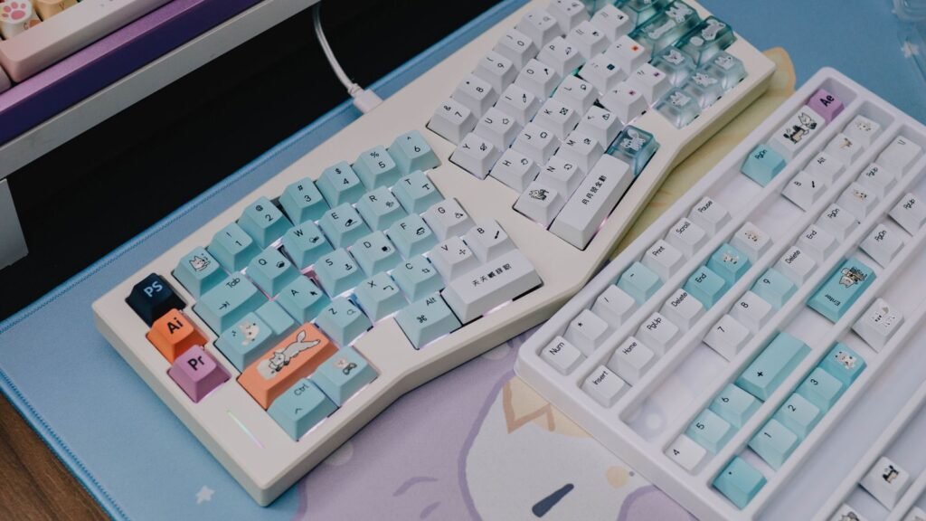 A cream colored split style mechanical keyboard with pastel blue keycaps and a white keyboard with matching keycaps on a cartoon desk mat