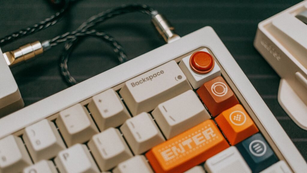 Close up of a cream mechanical keyboard with orange Enter key and a braided coiled cable with metal connectors on a dark desk