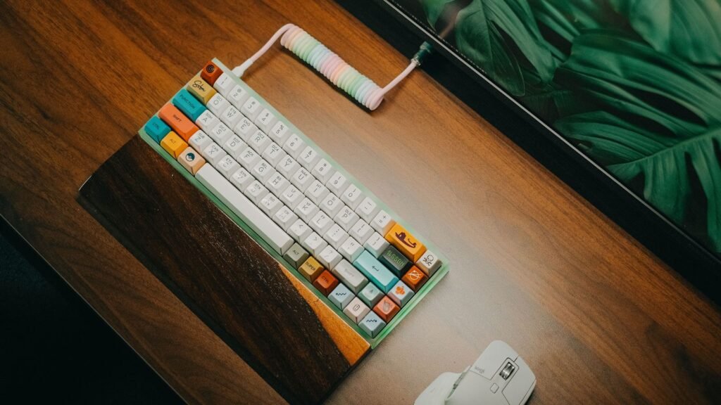 A small 60 percent style mechanical keyboard with colorful keycaps, a coiled cable and a wooden wrist rest on a wood desk