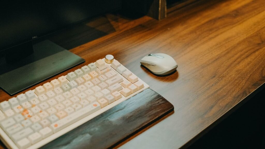 White compact mechanical keyboard with a silver knob and a dark wooden wrist rest on a wooden desk next to a white mouse and a monitor