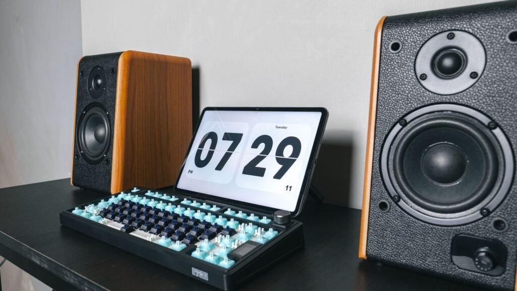 A black mechanical keyboard with most keycaps removed, exposing light blue and dark blue switches, in front of a tablet showing a clock between two wooden speakers