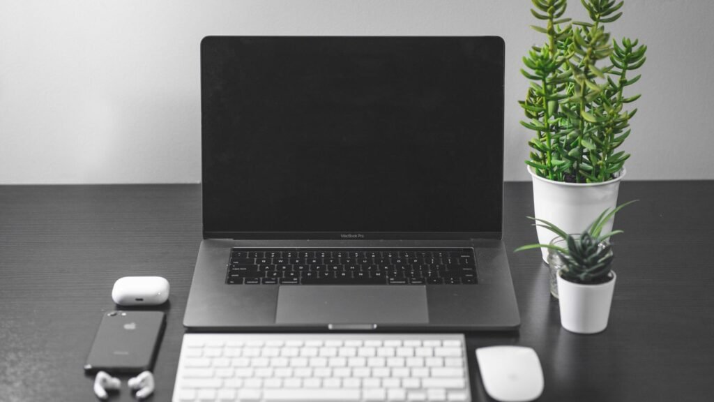 A MacBook Pro on a dark desk with a slim white Apple keyboard, white mouse, phone and potted plants