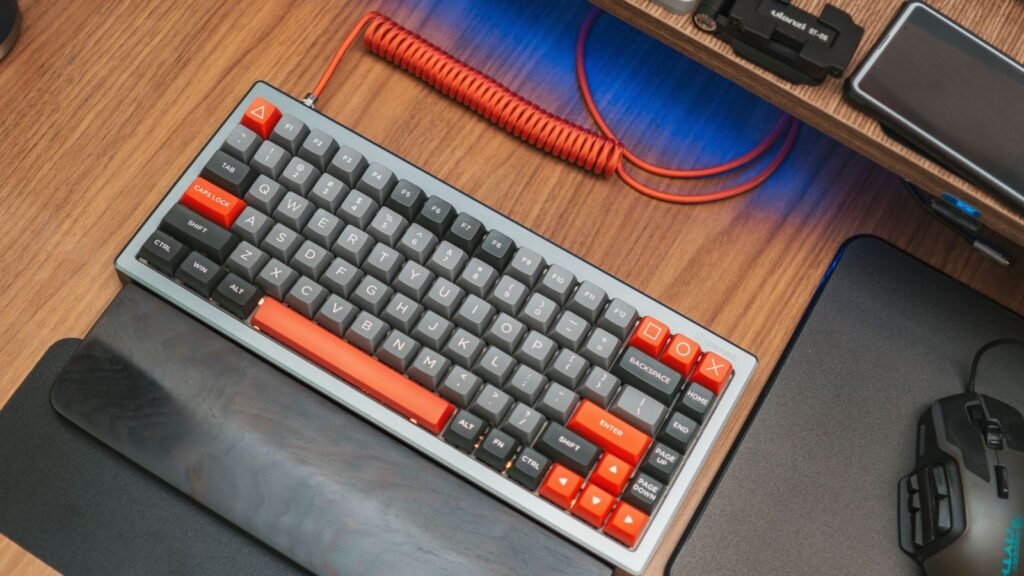 Compact mechanical keyboard with gray and orange keycaps, a coiled orange cable and a wrist rest on a wooden desk