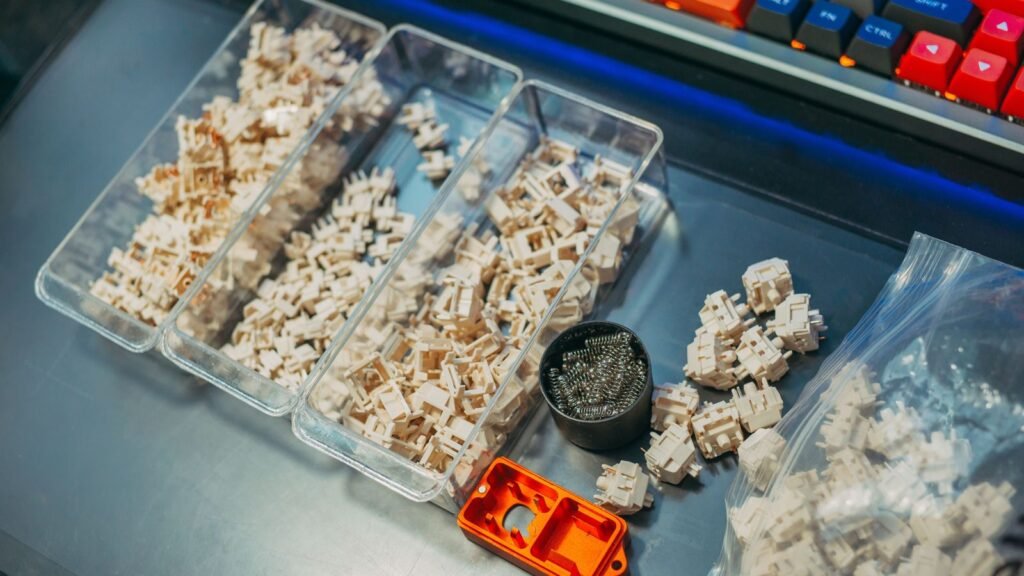 Clear trays full of cream colored keyboard switches next to a small cup of metal coil springs and loose switches on a desk