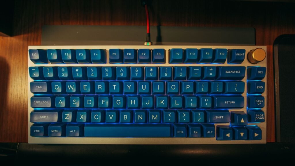 Top-down view of a silver 75 percent mechanical keyboard with blue keycaps, a full function row, a right-side column of navigation keys, an arrow cluster and a gold knob